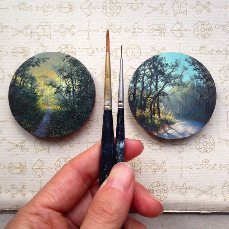 Hand holding fine paintbrushes between two detailed circular artworks, showcasing traditional Miniature Paintings in Lahore with scenic landscapes.