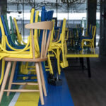 “Stacked colorful wooden chairs placed upside down on tables inside a closed modern café with glass walls.”